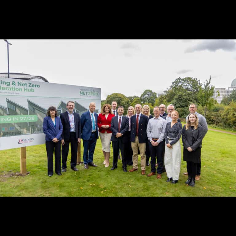 A group of thirteen people stand outdoors on grass beside a large sign for a Housing & Net Zero Acceleration Hub, with trees and a building in the background.