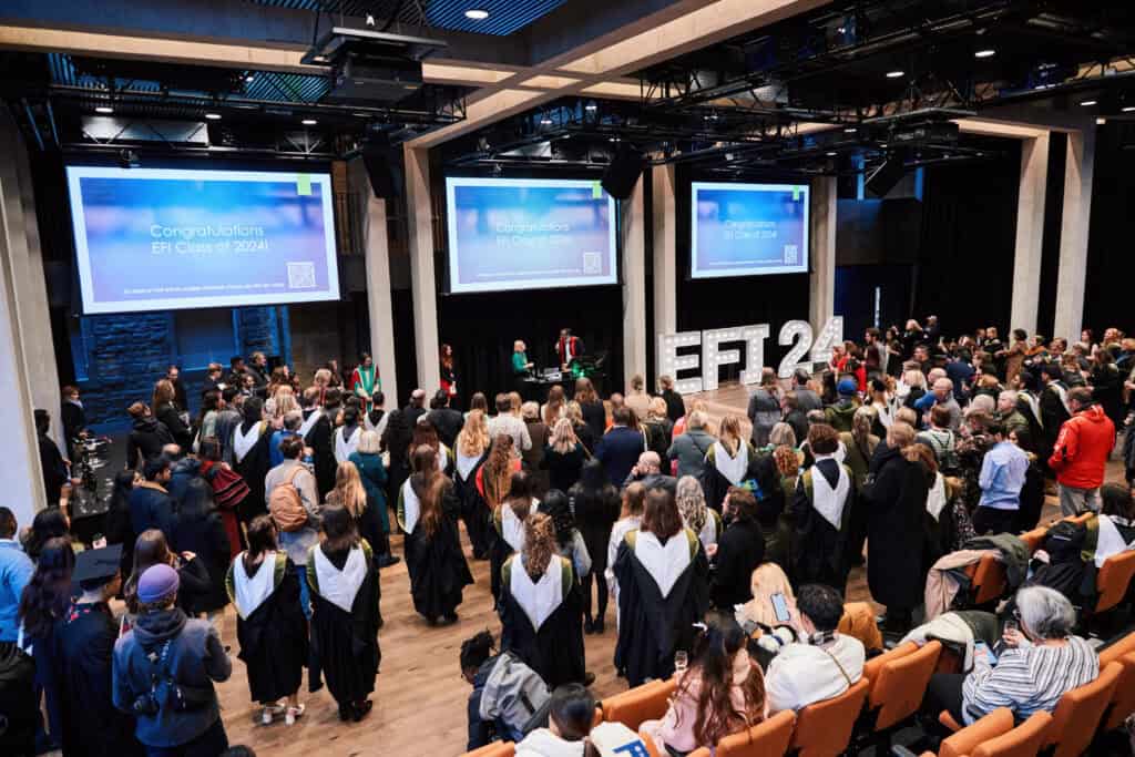 Graduates in caps and gowns face a stage during a graduation ceremony. Two screens display “Congratulations EFI Class of 2024.” Audience members are seated and standing in a modern auditorium.