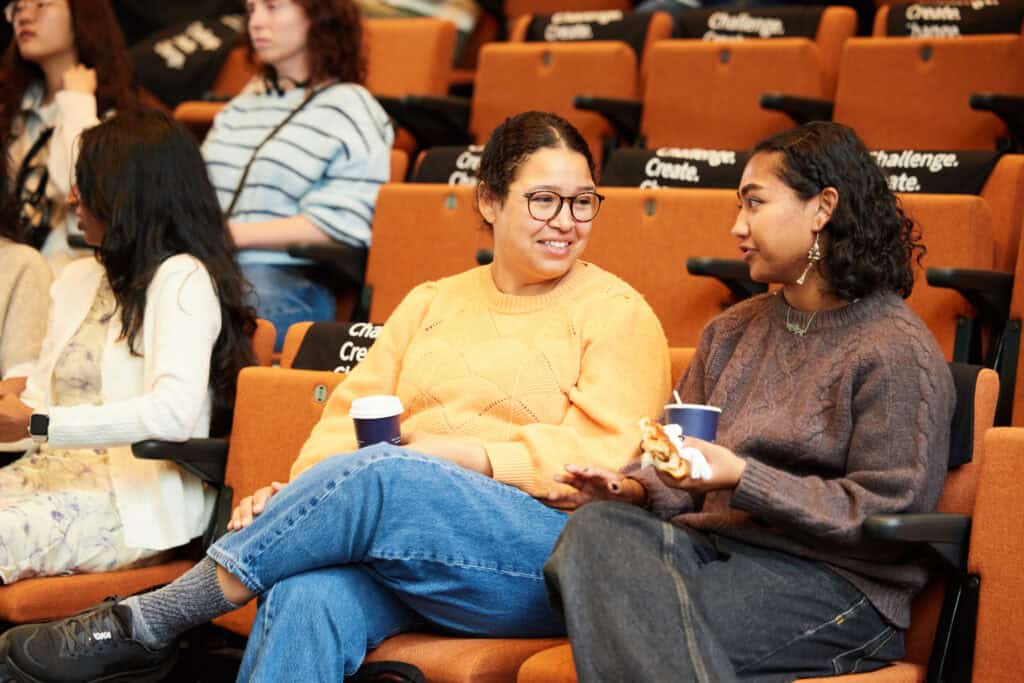 Two women sit and talk in an auditorium with orange seats. One wears a yellow jumper and glasses, holding a coffee cup; the other wears a brown jumper, holding a cup and snack. Others sit around them.
