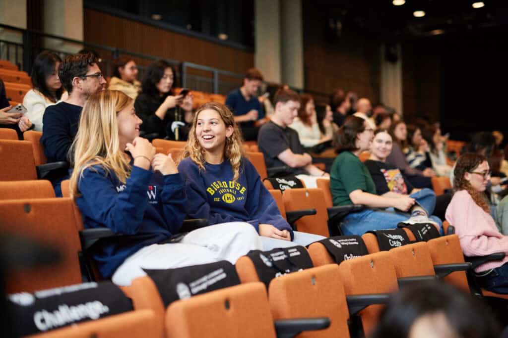 Students sit in an auditorium, some chatting and others looking at their mobiles. The seating is tiered with name tags visible on some chairs. The atmosphere appears casual and social.