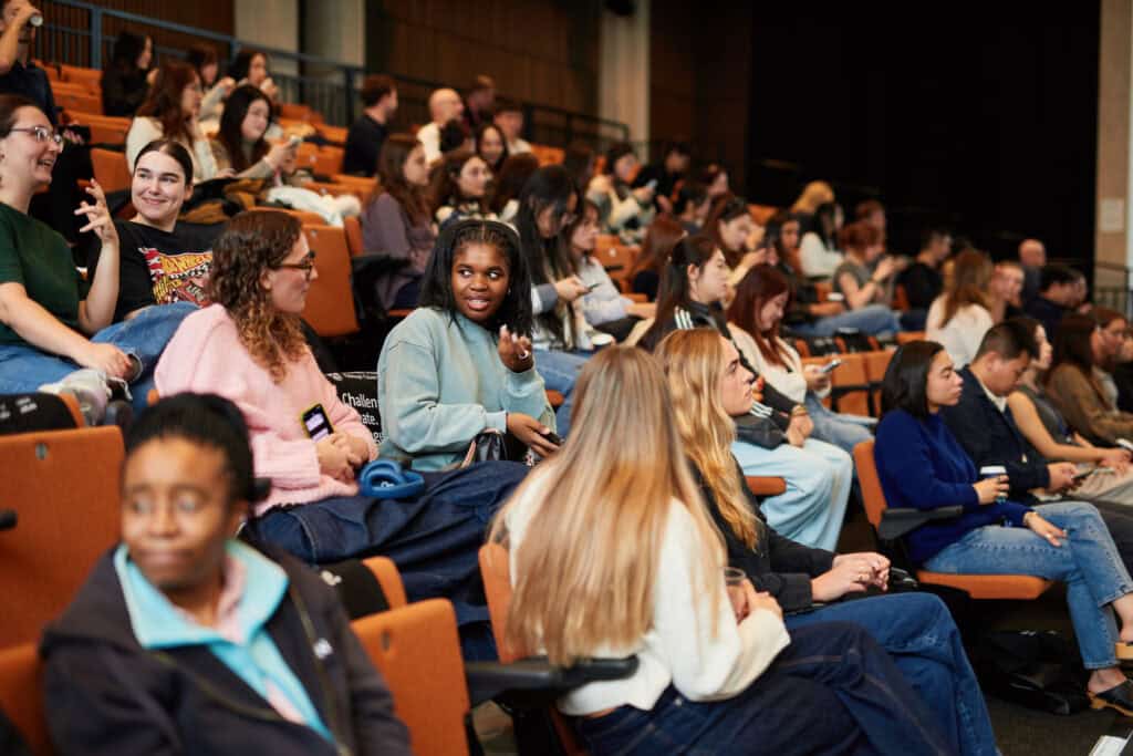 Students sit in an auditorium, some talking, others looking at mobiles or ahead. The room is brightly lit and filled with people, suggesting a class or event setting.