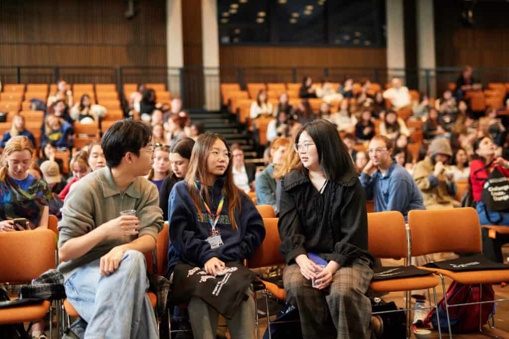 Three people sit and talk in the front row of an auditorium filled with people. The audience sits on orange chairs, and the background shows a modern space with large windows and wood panelling.