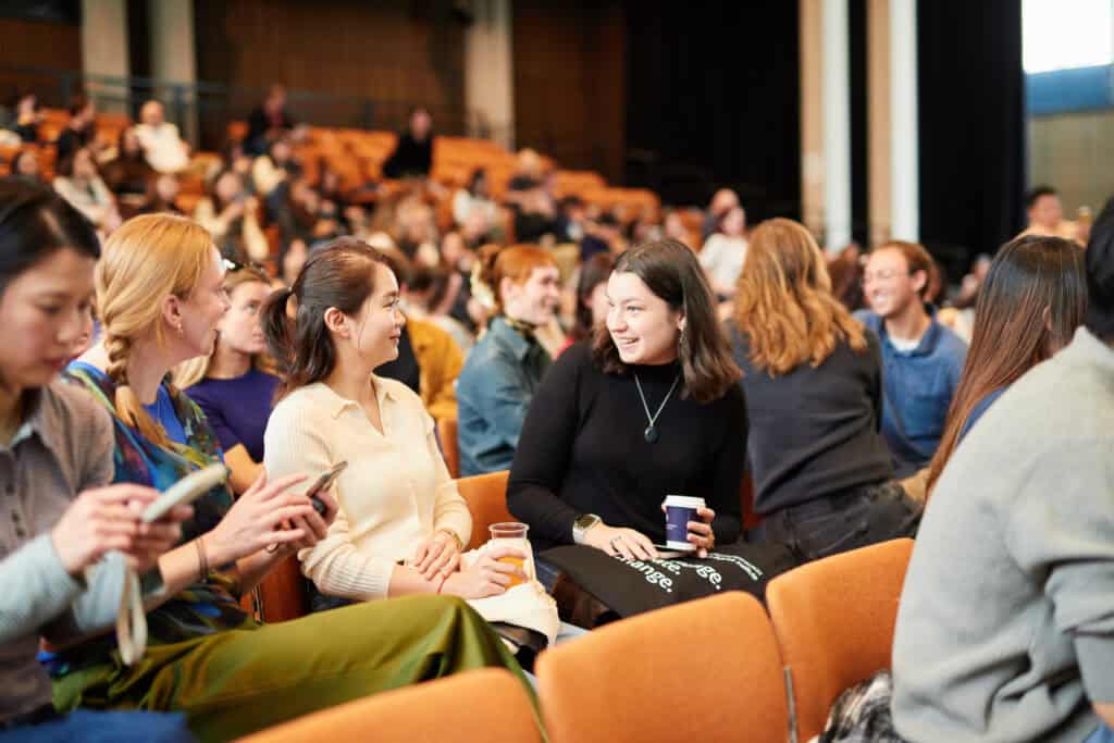 A group of people sit in an auditorium, talking and smiling before an event. Some hold drinks or programmes, while others chat. The room is filled with orange seats and natural light.