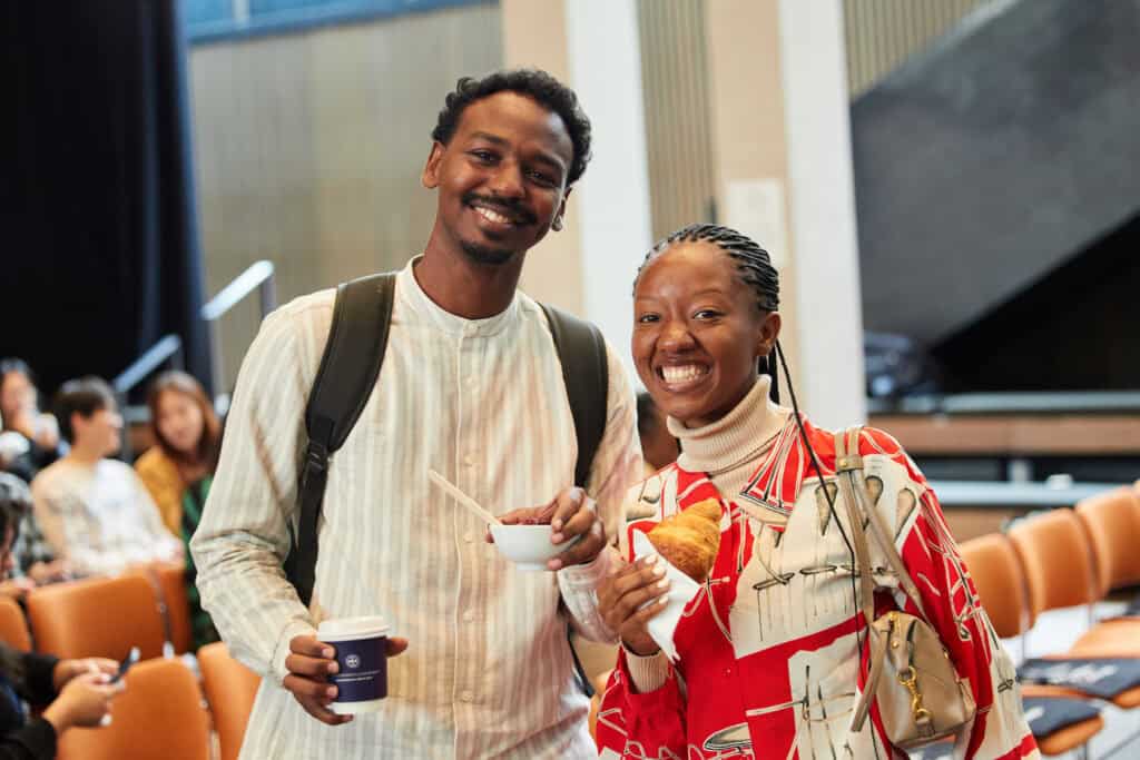 Two people smiling at the camera indoors, one holding a cup and bowl, the other holding a croissant. They appear to be at an event with empty chairs and other people seated in the background.