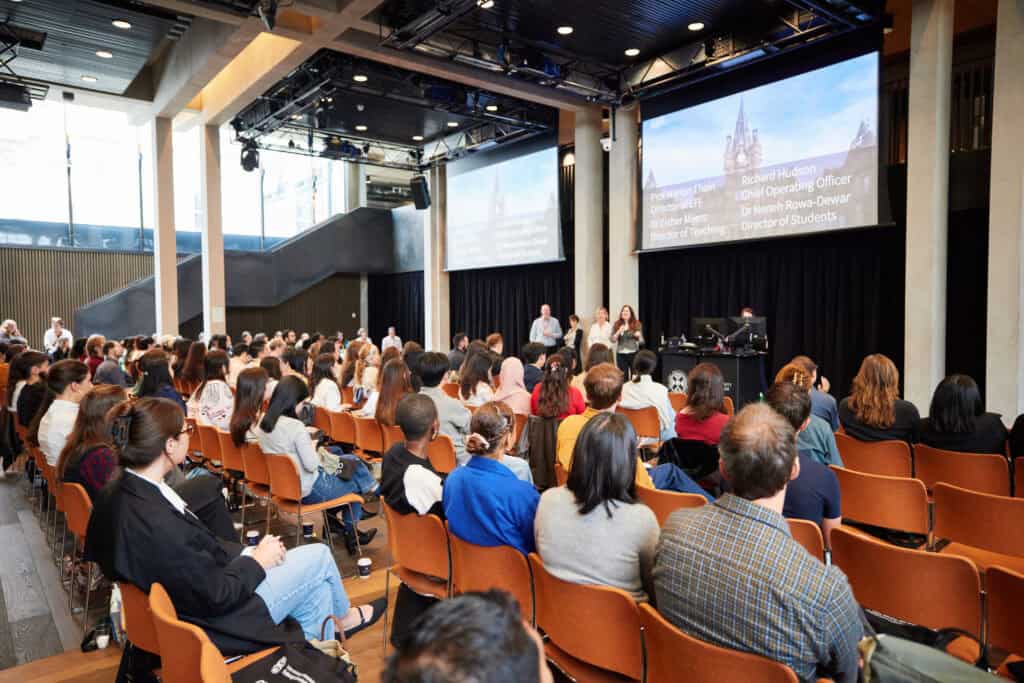 A large group of people sits in an auditorium, facing a stage with speakers and two large screens displaying presentation text. The room has modern design elements and natural lighting from large windows.