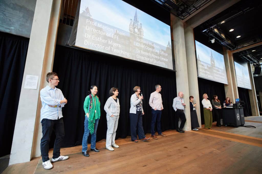 Seven people stand on stage beneath projected names and titles, next to a black curtain. Two more people stand by a lectern at the right, facing the audience in a large auditorium with wooden floors and tall columns.
