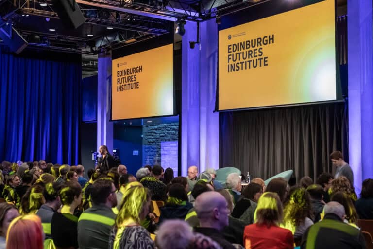 A large audience sits in an event space with two big screens displaying “Edinburgh Futures Institute.” Some people are standing near the stage, whilst others are seated, waiting for the event to begin.