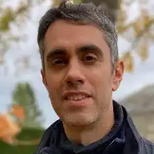 A man with short dark hair and some grey, wearing a dark jacket, stands outdoors with autumn trees and a blurred building in the background.